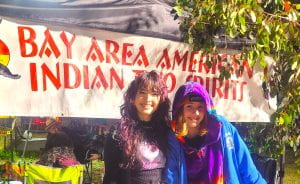 Joy's kid, Star Hagen-Esquerra (UCSC '22) and nibling Sequoyah Hagen smiling outside in front of a sign that reads Bay Area American Indian Two Spirits