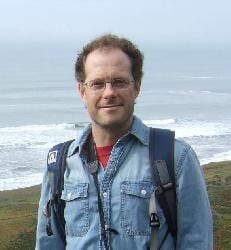 Image of Don Miller , wearing a denim shirt and backpack and standing in front of the ocean. 