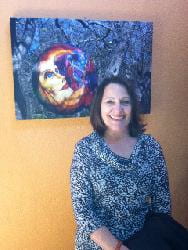 Photo of Gail smiling while standing in front of a mainly-blue piece of art and a yellow wall.