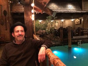 Photo of Mark with a black hat sitting with his arm resting on a railing that is overlooking a pond or pool. 